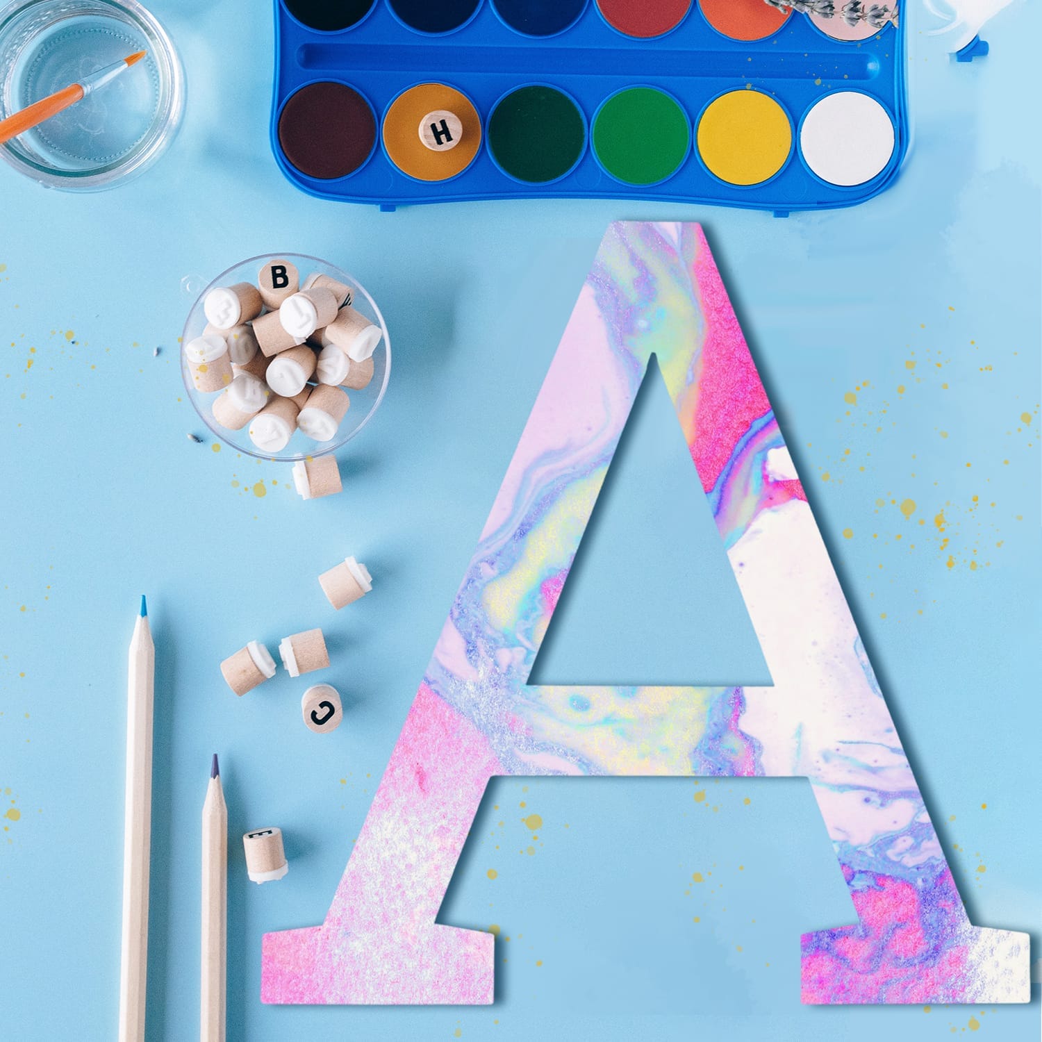 Pastel rainbow letter A surrounded by art supplies including watercolor paints, pencils, and alphabet stamps.